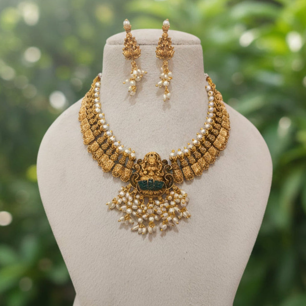 Gold and pearl jewelry set featuring a choker necklace with a central deity pendant, matching drop earrings, and a cream-colored display bust.