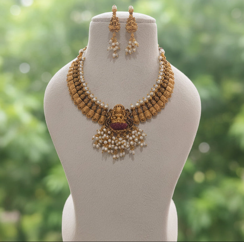 Gold and pearl traditional Indian jewelry set including a necklace, earrings, and a matching ring displayed on a beige jewelry bust.