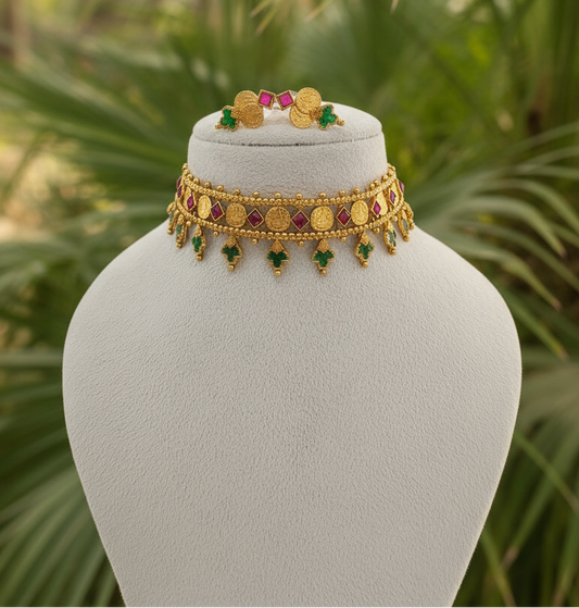 Gold-toned traditional Indian jewelry set displayed on a white jewelry bust, featuring a choker necklace with coin motifs, dangling green and red stones, and a matching ring, set against a blurred green foliage background.